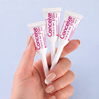 A hand with light pink manicured nails holds three white concealer tubes with pink and purple branding against a soft light blue background. The tubes are labeled 'Conceine Plus' and '12m'.
