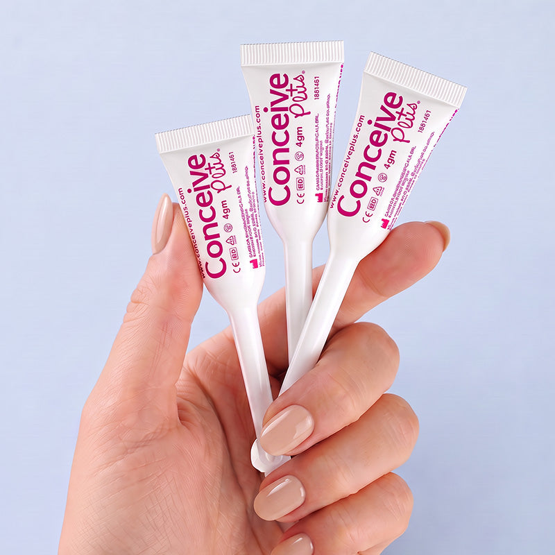 A hand with light pink manicured nails holds three white concealer tubes with pink and purple branding against a soft light blue background. The tubes are labeled 'Conceine Plus' and '12m'.